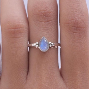 Könnte beinhalten: Ein Silberring mit einem birnenförmigen, irisierenden Mondstein-Cabochon in einer glatten Fassung. Die Ringschiene ist mit kleinen dekorativen Metallkugeln auf beiden Seiten des Mittelsteins verziert. Der Text LuxurySilver925 ist auf dem Ring zu sehen.