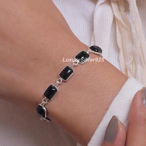 Peut inclure: Un bracelet à chaîne en argent composé de cinq pierres d'onyx noir rectangulaires serties dans des montures en argent poli. Le bracelet est relié par de petits anneaux en argent et porte le texte LuxurySilver925 imprimé en blanc près des pierres.