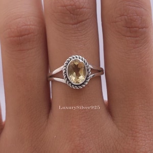 Könnte beinhalten: Ein Ring aus Sterlingsilber mit einem oval geschliffenen, blassgelben Citrin, der von einer gedrehten Kordelbordüre umrahmt wird. Die Ringschiene teilt sich in zwei dünne, polierte Silberstränge, die am Stein zusammenlaufen. Text LuxurySilver925 sichtbar.