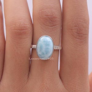 May include: A sterling silver ring featuring a smooth, oval-shaped, light blue Larimar gemstone set in a bezel. The band has a textured, twisted rope design. The text LuxurySilver925 is visible on the metal surface near the gemstone setting.