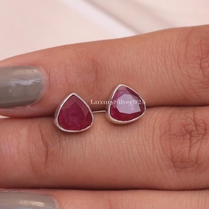 Könnte beinhalten: Ein Paar Ohrstecker mit facettierten, birnenförmigen roten Rubin-Edelsteinen in Silberfassungen. Der Text LuxurySilver925 ist zwischen den beiden Ohrringen zu sehen. Jeder Ohrring hat einen glatten, polierten Silberrand um den Stein.