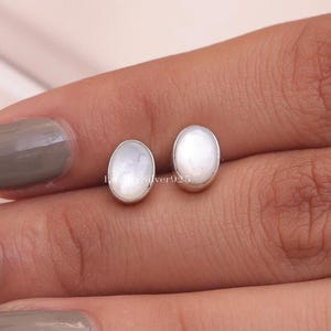 May include: A pair of oval stud earrings with a pearly white appearance, set in silver metal. The earrings are displayed on a person's hand. The earrings are approximately 1 cm in length.