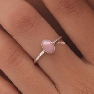 Op de afbeelding: Een zilveren ring met een lichtroze ovale edelsteen. De ring heeft een dunne band en een eenvoudig ontwerp. De edelsteen is gezet in een bezel setting. De ring wordt op een vinger getoond.