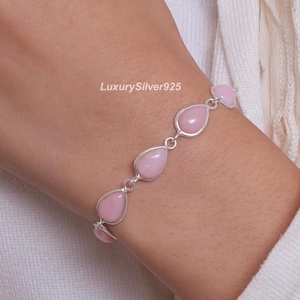 Peut inclure: Un bracelet en chaîne argentée composé de cinq pierres lisses rose clair en forme de goutte d'eau reliées entre elles. Le texte LuxurySilver925 est visible près du centre du bracelet. Le design présente des montures en métal poli.
