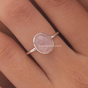 Könnte beinhalten: Ein silberner Ring mit einem ovalen, hellrosa Edelstein. Der Ring hat ein schlichtes, elegantes Design. Der Text "LuxurySilver925" ist auf dem Ring sichtbar. Dieser Ring ist ein Schmuckstück.