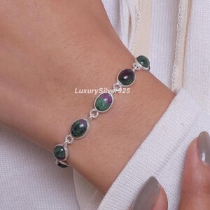 Könnte beinhalten: Ein Silberarmband mit einer Reihe ovaler, im Cabochon-Schliff gefertigter Rubin-Zoisit-Edelsteine, die durch dünne silberne Metallglieder verbunden sind. Die Steine weisen ein gesprenkeltes Muster aus tiefgrünen und dunkelroten Farbtönen auf.
