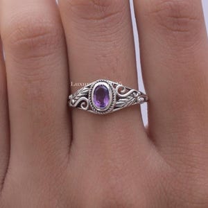 Könnte beinhalten: Ein Silberring mit einem ovalen, violetten Amethyst-Edelstein in einer seilartig texturierten Fassung. Die Ringschiene zeigt filigrane Metallarbeiten mit geschwungenen blattartigen Mustern, die sich beidseitig von der Fassung erstrecken.