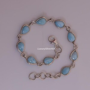 May include: A silver chain bracelet featuring eight teardrop-shaped light blue stones set in metal bezels. The bracelet includes a lobster claw clasp and an adjustable extender chain. The text LuxurySilver925 is visible in the centre of the image.