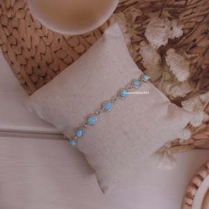 May include: A silver chain bracelet featuring multiple light blue, teardrop-shaped stones set in metal bezels. The bracelet is displayed on a small, square, off-white fabric pillow. The text LuxurySilver925 is printed in small white letters.