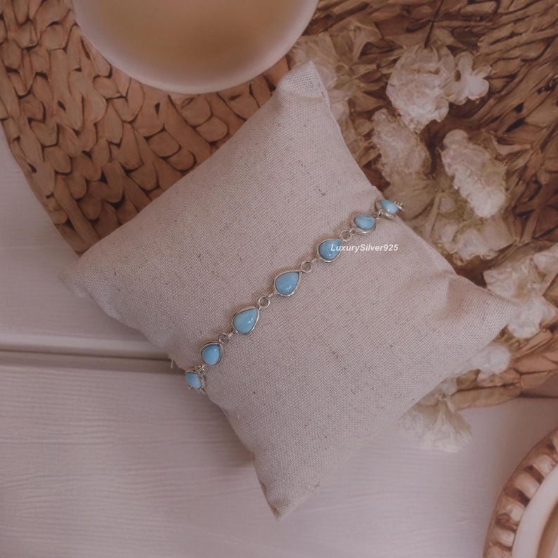 May include: A silver chain bracelet featuring multiple light blue, teardrop-shaped stones set in metal bezels. The bracelet is displayed on a small, square, off-white fabric pillow. The text LuxurySilver925 is printed in small white letters.