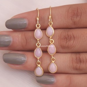 May include: A pair of gold-toned drop earrings, each featuring three vertically aligned, teardrop-shaped light pink stones set in metal frames. The earrings hang from simple hook wires and display a delicate, linear design with smooth, polished surfaces.