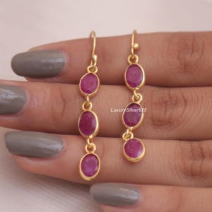 Pode incluir: Um par de brincos pendentes em tom dourado, cada um com três pedras ovais de cor rosa magenta alinhadas verticalmente. Os brincos estão suspensos por ganchos dourados simples. O texto LuxurySilver925 é visível no centro da imagem.