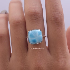 May include: A silver ring featuring a square, polished, light blue Larimar gemstone with white marbling patterns. The stone is set in a smooth silver bezel. The text LuxurySilver925 is visible in small font below the stone on the silver band.