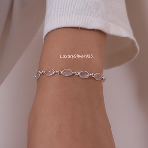 May include: A silver chain bracelet featuring a series of oval-shaped, translucent light pink stones set in polished metal bezels. The text LuxurySilver925 is visible above the bracelet. The design consists of repeating links connecting each stone.