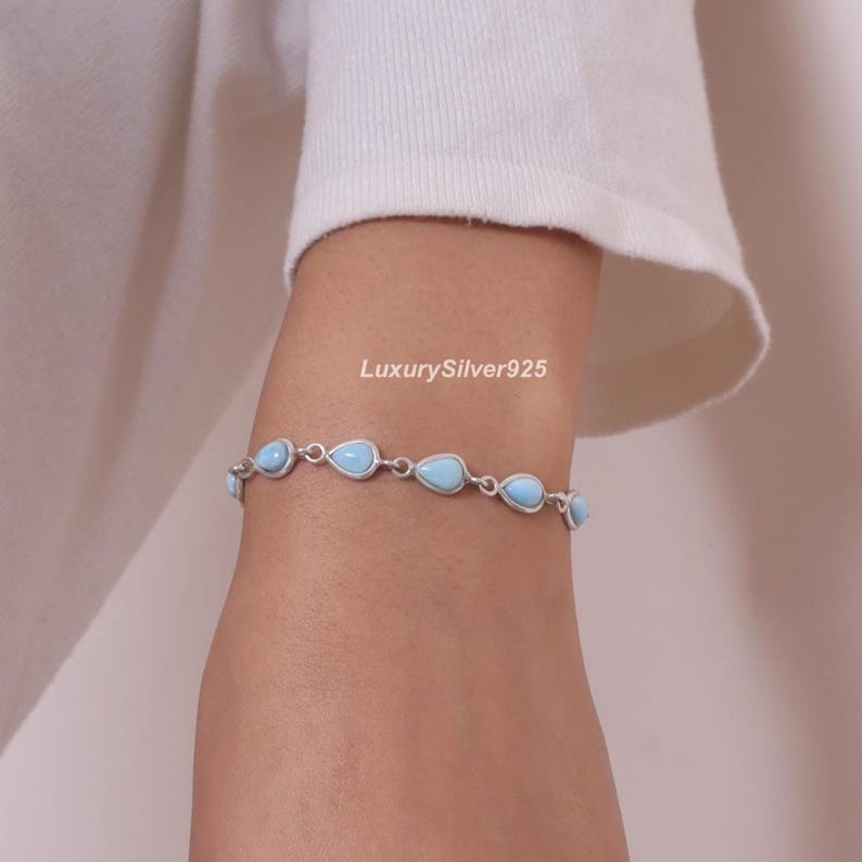May include: A silver chain bracelet featuring a series of light blue, teardrop-shaped stones set in metal frames. The bracelet is displayed on a wrist, with the text LuxurySilver925 visible above the jewellery piece, which consists of multiple links.
