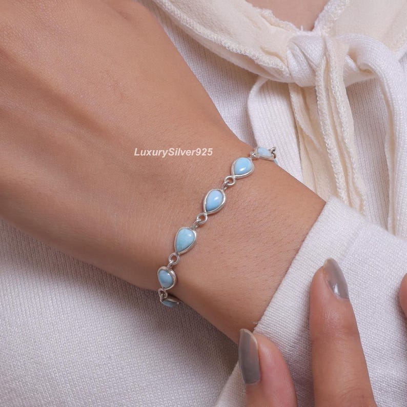 May include: A silver chain bracelet featuring multiple teardrop-shaped light blue stones set in metal bezels. The bracelet is delicate and links the stones together in a repeating pattern. The text LuxurySilver925 is visible near the jewellery.