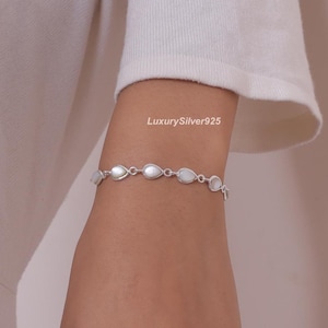 Könnte beinhalten: Ein silbernes Kettenarmband mit einer Reihe von tropfenförmigen weißen Perlmutt-Einlagen, die in polierte silberne Metallglieder gefasst sind. Das Armband wird am Handgelenk getragen, wobei der Text LuxurySilver925 im Hintergrund zu sehen ist.