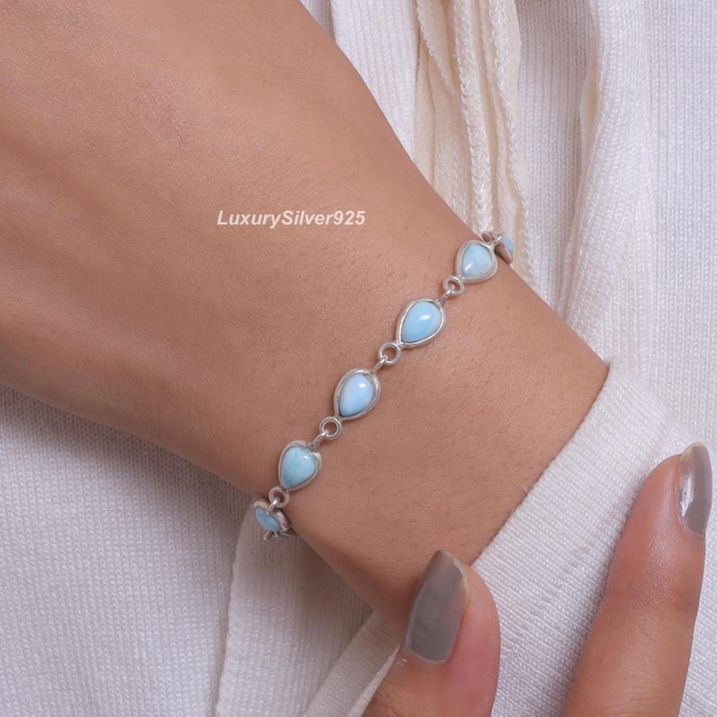 May include: A silver chain bracelet featuring multiple teardrop-shaped light blue stones set in metal bezels. The bracelet is linked together with small silver rings. The text LuxurySilver925 is visible near the bracelet on the skin.