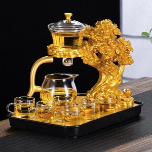 May include: A golden tea set with a tree-shaped stand, a glass teapot, and several small teacups. The set includes a glass tea infuser and a black tray. The tea is a light amber color.