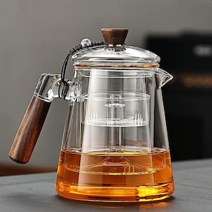 Puede incluir: Una tetera de vidrio transparente con asa y tapa de madera. La tetera tiene un infusor incorporado y está llena de líquido color ámbar. Un cordón negro con una cuenta decorativa está unido a la tapa. La tetera está diseñada para preparar té.