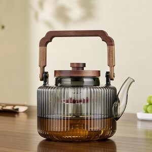Puede incluir: Una tetera de vidrio transparente con asa y tapa de madera oscura. La tetera tiene un diseño acanalado y contiene té. El asa es arqueada y está unida al cuerpo de vidrio con bisagras metálicas.