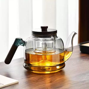 May include: A clear glass teapot with a dark brown handle and lid. The teapot is filled with amber-colored liquid, likely tea, and has a built-in infuser. The teapot sits on a wooden surface, suggesting a tea-drinking setting.