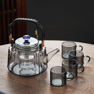 Puede incluir: Una tetera de vidrio transparente con asa negra y pomo azul, acompañada de cuatro tazas de vidrio a juego. La tetera tiene un diseño único y está sobre una superficie de madera.