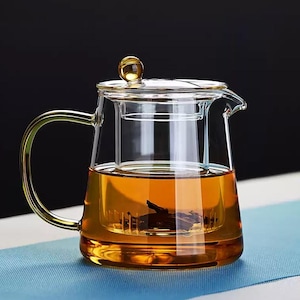 Puede incluir: Una tetera de vidrio transparente con asa y tapa doradas. La tetera tiene un infusor incorporado y está llena de té de color ámbar. El asa y el pomo de la tapa tienen un acabado dorado. La tetera está sobre una superficie azul.