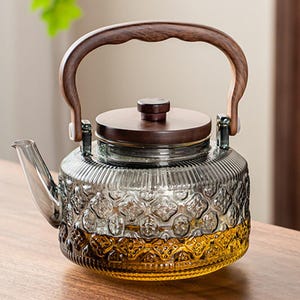 Op de afbeelding: Een glazen theepot met een houten handvat en deksel. De heldere glazen body heeft een decoratief patroon en een kleurovergang, van helder naar amber. Het handvat is gebogen en het deksel heeft een kleine knop.