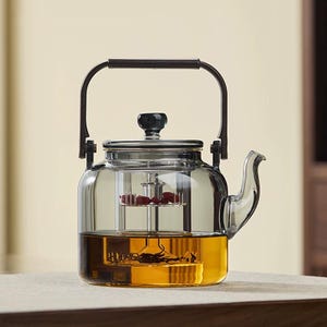 May include: A clear glass teapot with a dark handle and lid. The teapot has a built-in infuser and is filled with tea and red berries. The teapot is designed for brewing tea and has a spout for pouring.