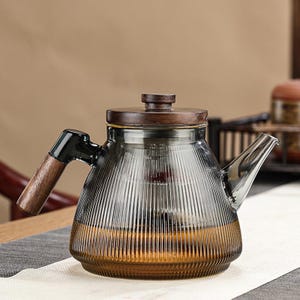 Peut inclure: Théière en verre avec une poignée et un couvercle en bois. Le corps en verre transparent présente des rainures verticales et une légère teinte ambrée à la base. La poignée et le couvercle sont en bois foncé. La théière est conçue pour infuser le thé.