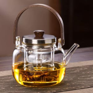 May include: A clear glass teapot with a wooden handle and lid. The teapot is filled with amber-colored liquid, likely tea, and has a built-in infuser. The handle is arched and made of wood, and the lid is silver with a wooden knob.