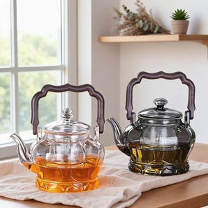 May include: Two glass teapots with dark brown wooden handles and lids. One teapot is clear glass filled with amber liquid, and the other is a dark grey tinted glass. Both feature a curved spout and a decorative base with a scalloped edge design.