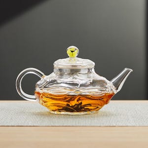 May include: A clear glass teapot with a wavy design and a yellow and green knob on the lid. The teapot is filled with amber-colored tea. The handle and spout are curved. The teapot is sitting on a light gray surface.