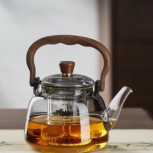 Puede incluir: Una tetera de vidrio transparente con asa y tapa de madera. La tetera tiene un infusor incorporado y está llena de líquido color ámbar. El asa es curva y de madera oscura. La tapa también es de madera con un acento de latón.