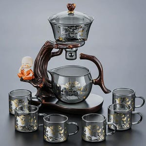 May include: A glass tea set with a wooden stand. The set includes a teapot, a serving pot, a tea strainer, and six teacups. The glass pieces are decorated with gold-coloured horse designs. A small statue of a monk is on the side.