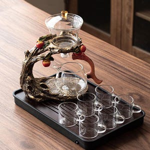 Può includere: Un set da tè con un supporto decorativo a forma di albero, una teiera in vetro, un infusore in vetro e sei piccole tazze di vetro. Il supporto ha una finitura marrone con accenti di mela rossa. Il set è su un vassoio nero.