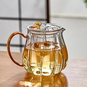 May include: A clear glass teapot with a curved handle and a lid. The teapot is filled with a golden-coloured liquid, possibly tea. The handle and lid accents have a golden hue. The teapot is designed for brewing tea.