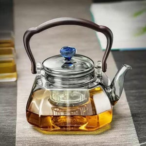 Könnte beinhalten: Eine transparente Glas-Teekanne mit einem braunen Griff und einem blauen Knopf auf dem Deckel. Die Teekanne ist mit bernsteinfarbener Flüssigkeit gefüllt, wahrscheinlich Tee. Das Design ist modern und elegant, geeignet zum Servieren von Tee.