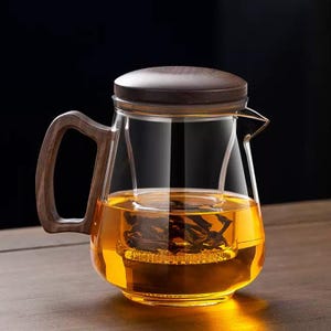Op de afbeelding: Een heldere glazen theepot met een houten handvat en deksel. De theepot is gevuld met amberkleurige thee en heeft een ingebouwde infuser. Het handvat en deksel zijn van donker hout. De theepot is ontworpen voor het zetten van thee.