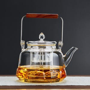 May include: A clear glass teapot with a stainless steel handle and a wooden grip. The teapot has a glass infuser inside and is filled with amber-colored liquid. The teapot is sitting on a wooden surface.