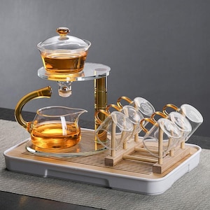 Op de afbeelding: Een glazen theeset met goudkleurige handvatten. De set bevat een theepot, een serveervaas en verschillende kleine kopjes, allemaal gerangschikt op een bamboe dienblad. De thee is amberkleurig.