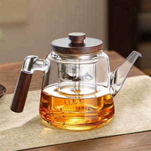 May include: A clear glass teapot with a wooden handle, lid, and infuser. The teapot is filled with amber-colored liquid. The handle and lid are made of dark wood. The teapot is sitting on a light-colored surface.