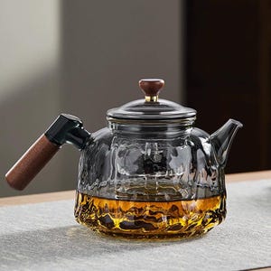 May include: A clear glass teapot with a wooden handle and lid. The teapot is filled with a golden-brown liquid, likely tea. The handle is a dark color with a wooden grip. The lid has a wooden knob.
