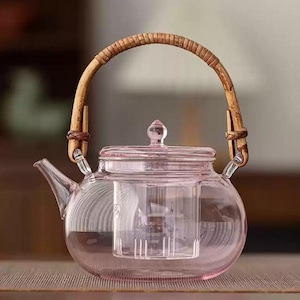 Könnte beinhalten: Eine klare Glas-Teekanne mit rosa Farbton, mit rundem Körper und Bambusgriff. Die Teekanne enthält einen Glas-Teefilter und einen Deckel mit kleinem Knopf. Der Griff ist mit braunem Bindfaden befestigt.