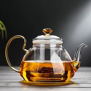 May include: A clear glass teapot with a gold handle and spout. The teapot is filled with amber-colored tea and has a glass lid with a decorative amber-colored knob. The teapot sits on a clear glass base.