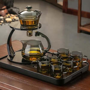 Può includere: Un set da tè in vetro con una teiera, una brocca, un coperchio e sei tazzine. Il set è esposto su un vassoio nero con un supporto nero. Il tè è color ambra.