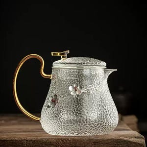 Könnte beinhalten: Eine weiße Keramik-Tasse mit schwarzem Henkel und dem Text "But First, Coffee" in fetter, schwarzer Schrift. Die Tasse steht vor einem verschwommenen Hintergrund, der eine gemütliche Umgebung suggeriert.