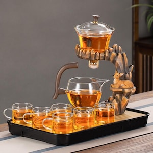 Puede incluir: Un juego de té con una tetera y tazas de vidrio transparente, llenas de líquido ámbar. El juego incluye un soporte decorativo con una figura de ciervo y una bandeja. El juego está diseñado para servir té.