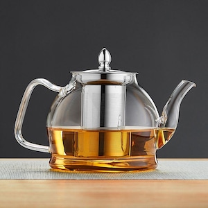 Może przedstawiać: Przezroczysty szklany czajniczek z zaparzaczem i pokrywką ze stali nierdzewnej. Czajniczek jest wypełniony bursztynowym płynem, prawdopodobnie herbatą, i ma zakrzywiony uchwyt i dzióbek. Czajniczek stoi na jasnej powierzchni na ciemnym tle.